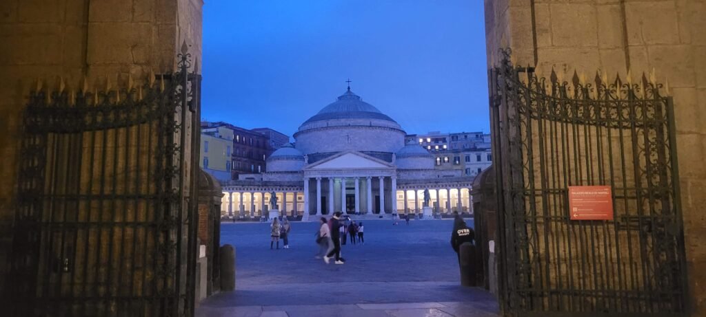 Piazza del Plebiscito widoczny z wejścia do Zamku Królewskiego w Neapolu. Zdjęcie zrobione wczesnym wieczorem - widoczna na drugim planie Bazylika jest już oświetlona, ale niebo wciąż jest niebieskie,