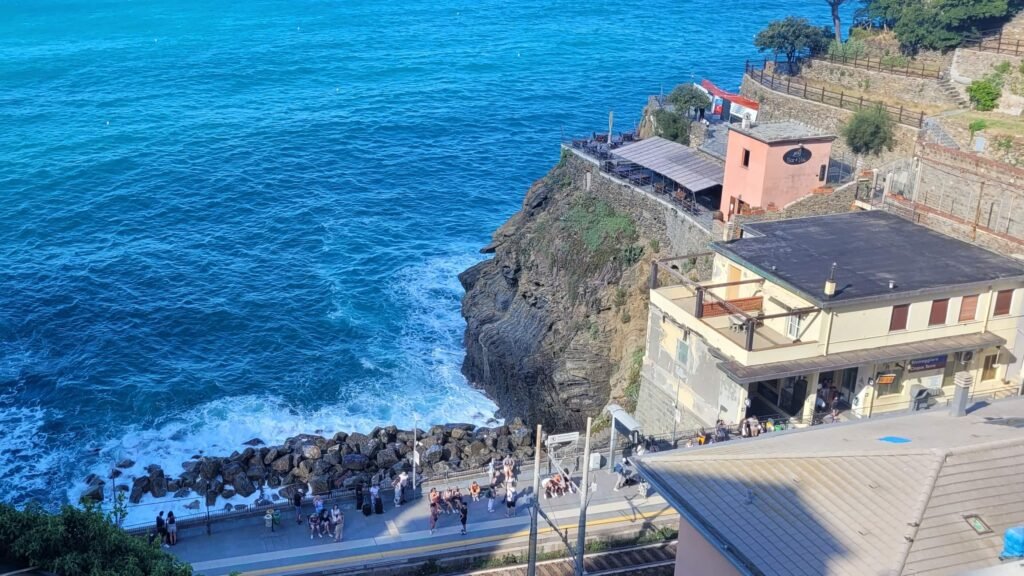 Widok na stację kolejową w Riomaggiore (Cinque Terre). Stacja położona jest na skale bezpośrednio nad lazurowym morzem. Pociągi są najwygodniejszym sposobem na przemieszczanie się pomiędzy miasteczkami. Zdjęcie zrobione z góry. Na stacji widać malutkie sylwetki ludzi.