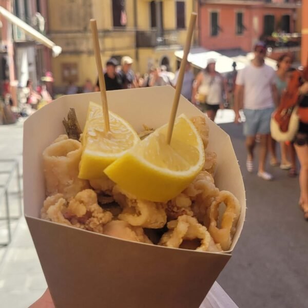 Na zdjęciu widoczne jest Fritto Misto - rożek zawierający smażone owoce morza,  w które wbite zostały dwa patyczki do szaszłyków, na które nadziane grube plastry cytryny. Rożek widoczny jest na tle uliczki w Riomaggiore z kolorowymi budynkami. W tle widać nieostre sylwetki tłumu ludzi.