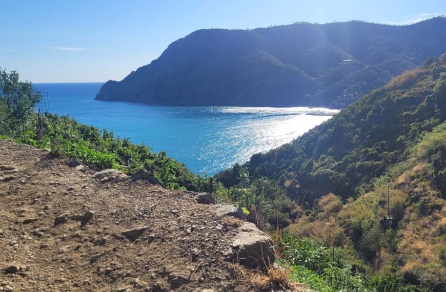 Chcąc się przejść po Błękitnym Szlaku (Azure Trail), potrzebujesz kupić Cinque Terre Card. Na zdjęciu przedstawiono krajobraz widziany z trasy pomiędzy Monterosso a Vernazzą. Na pierwszym planie z lewej strony widać fragment szlaku: szarobrązowy piasek i kamienie, nieco dalej z prawej strony widoczne jest wzgórze porośnięte ciemnozielonymi roślinami. Jeszcze dalej, pomiędzy nimi widać błękitne morze z padającymi na nie promieniami jasnego słońca. W tle widoczne są ciemnozielone, niemal czarne wzgórza, schodzące do morza.