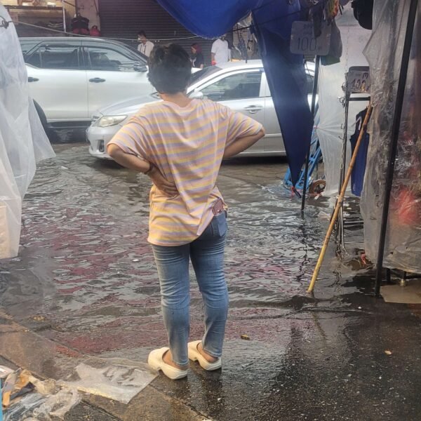 Ulica Bangkoku po krótkiej ulewie. W centrum stoi kobieta ubrana w białą koszulę i jeansy, patrzy na wodę z rękami opartymi na biodrach. Po drugiej stronie ulicy stoją samochody z kołami zanurzonymi w wodzie.