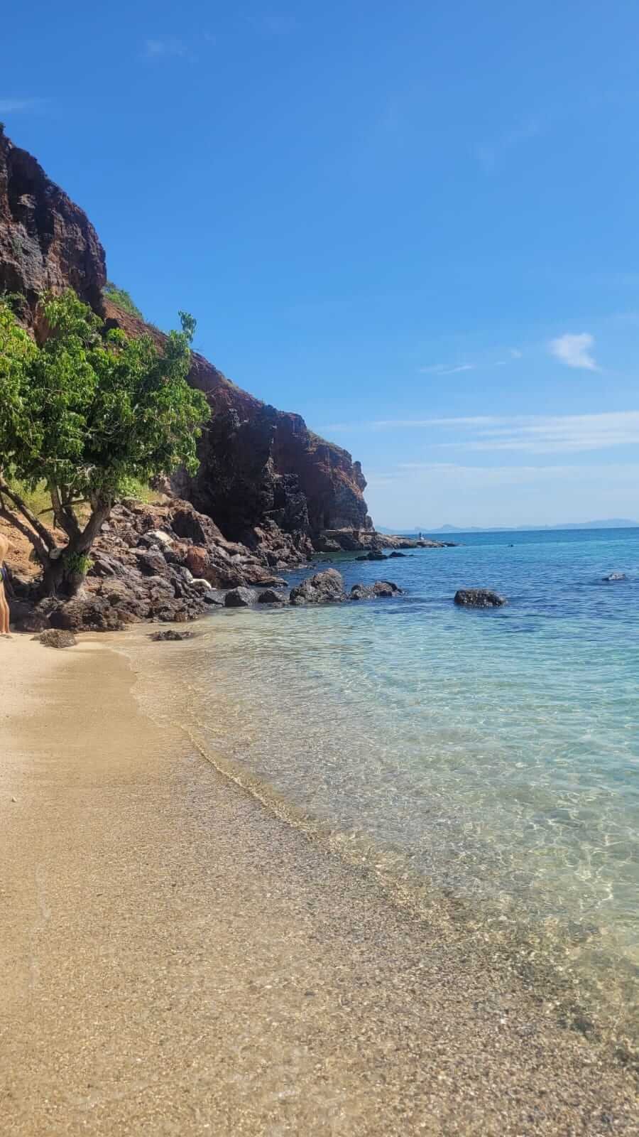 Tajlandia słynie z pięknych plaż. Wybrzeże - krystalicznie czysta błękitna woda obmywa plażę ze złotym (jasnopomarańczowym) piaskiem. Na końcu plaży widać szare kamienie przy klifie i samotne drzewo z zieloną koroną.